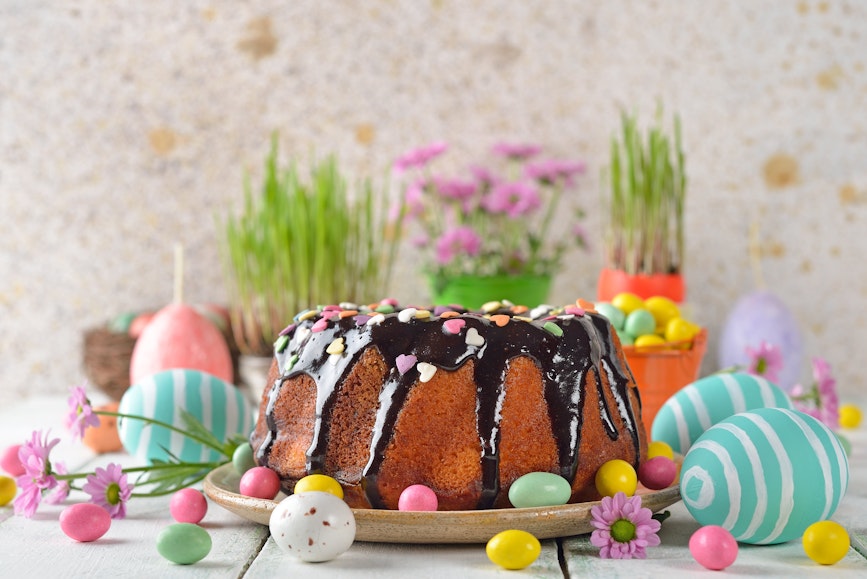 Easter Goods white wooden table topped with easter egg decorations and a giant sponge cake drizzled with chocolate and mini easter eggs in front of pots of flowers