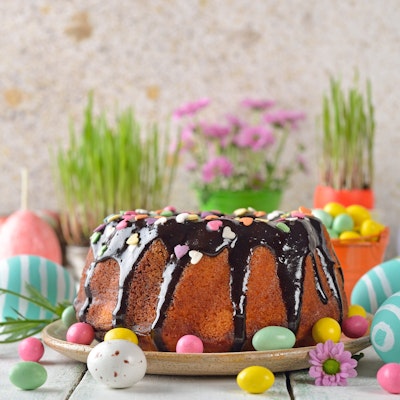 white wooden table topped with easter egg decorations and a giant sponge cake drizzled with chocolate and mini easter eggs in front of pots of flowers 