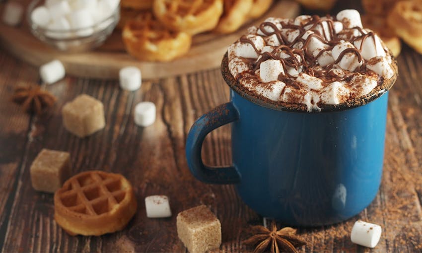 Blue mug of hot chocolate topped with marshmallows, caramel drizzle on a wooden table with mini waffles, sugar cubes and marshmallows 