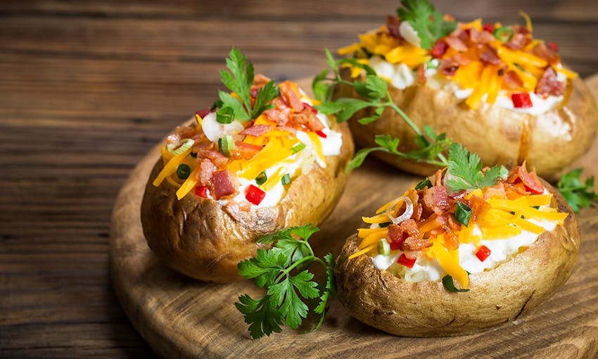 Three crispy baked potatoes topped with cream, cheddar, bacon and fresh spring onion on a wooden serving board 
