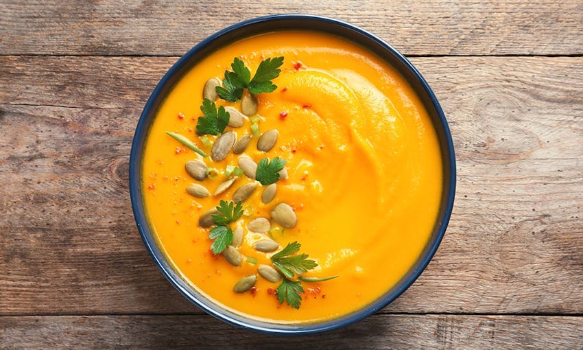 A bowl of creamy pumpkin soup topped with with pumpkin seeds and herbs set on a wooden table 
