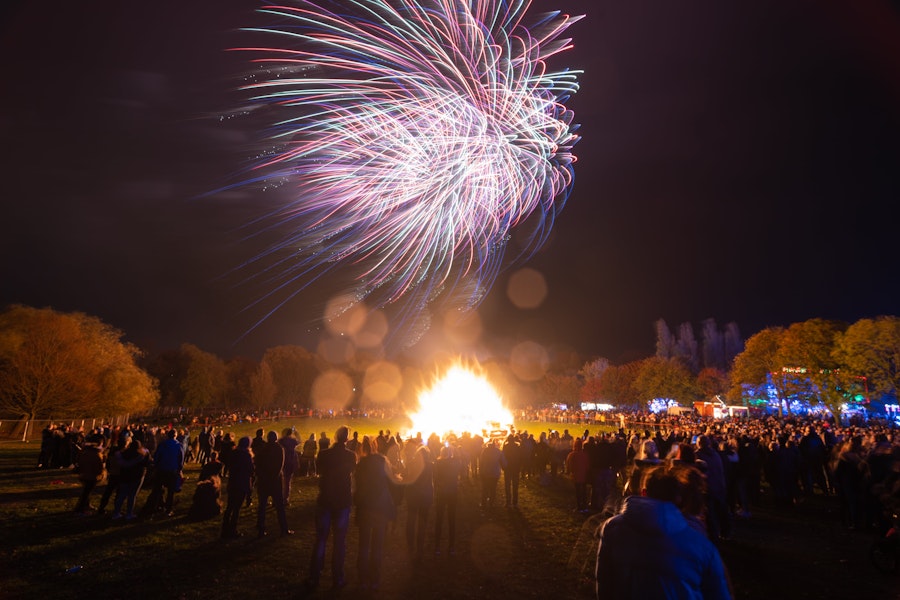Crowds gather around large bonfire surrounded by autumnal trees with fireworks lighting up the sky 