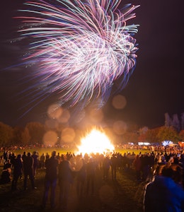 Crowds gather around large bonfire surrounded by autumnal trees with fireworks lighting up the sky 