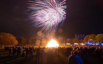 Crowds gather around large bonfire surrounded by autumnal trees with fireworks lighting up the sky 