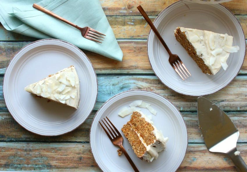 Three slices of carrot cake with frosting laid out on wooden table with light blue tablecloth and cutlery 