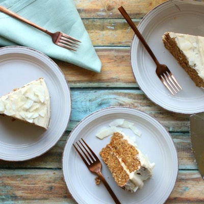 Three slices of carrot cake with frosting laid out on wooden table with light blue tablecloth and cutlery 