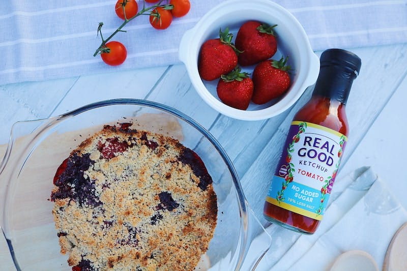 Real Good Ketchup’s Red Berry Cobbler Berry cobbler in clear baking dish next to a bottle of real good ketchup and bowl of strawberries