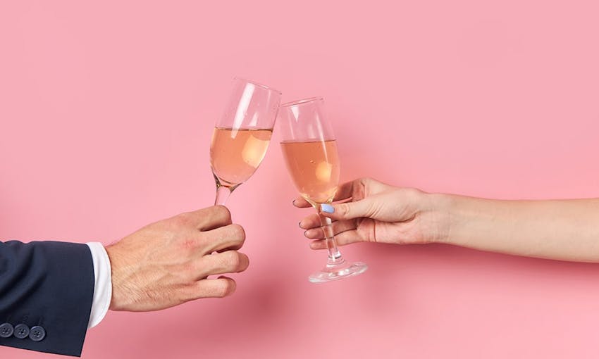 the arms of a man in a suit and a lady clinking two prosecco glasses filled with sparkling tea in front of a pink background  