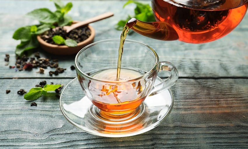 hot herbal tea in a clear glass cup and saucer with hot tea being poured in from a teapot next to a small dish of tea leaves