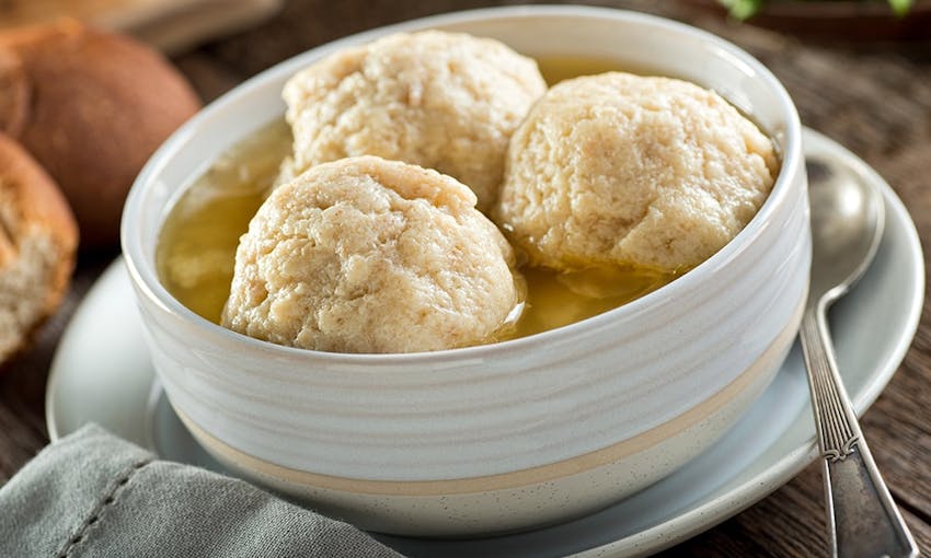 three light and fluffy matzo balls in chicken soup in a small white bowl with a soup spoon 