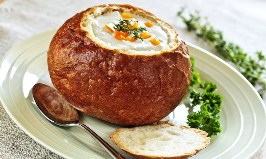 crusty sourdough bread with top cut off and filled with soup served with kale on a white plate and silver soup spoon 