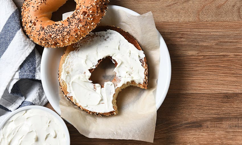 Brown seeded bagel halved with a cream cheese filling served on a white plate next to a striped white and navy tea towel 