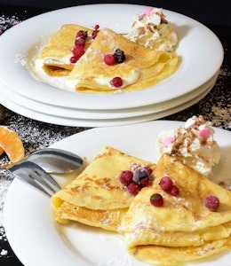 two plates of sweet crepes on a decorated table dusted with icing sugar and topped with berries 