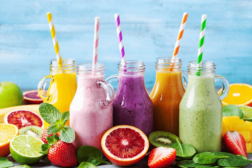 yellow, pink, purple, orange and green smoothies in glass jars with striped spoons surrounded by fresh fruit 