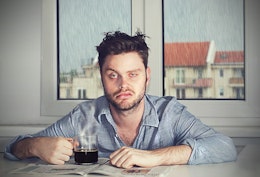 a tired hungover man with a defeated expression sat infront of a window where it is raining outside with a black coffee 