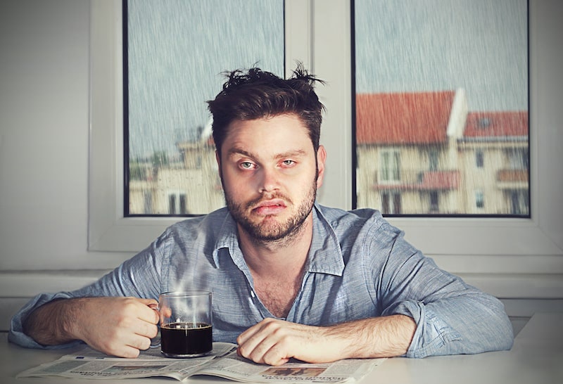 a tired hungover man with a defeated expression sat infront of a window where it is raining outside with a black coffee 