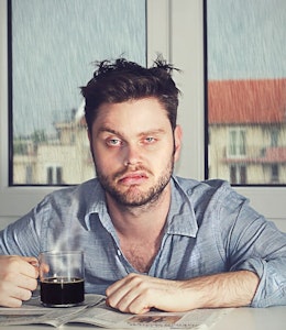 a tired hungover man with a defeated expression sat infront of a window where it is raining outside with a black coffee 