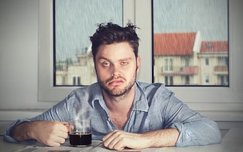 How to stop yourself feeling like this? Read on to find out. a tired hungover man with a defeated expression sat infront of a window where it is raining outside with a black coffee