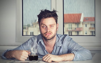 a tired hungover man with a defeated expression sat infront of a window where it is raining outside with a black coffee 