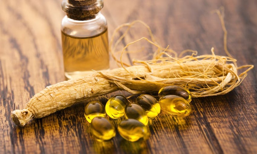 Red ginseng oil in a small glass corked bottle next to ginseng capsules and a piece of ginseng plant 