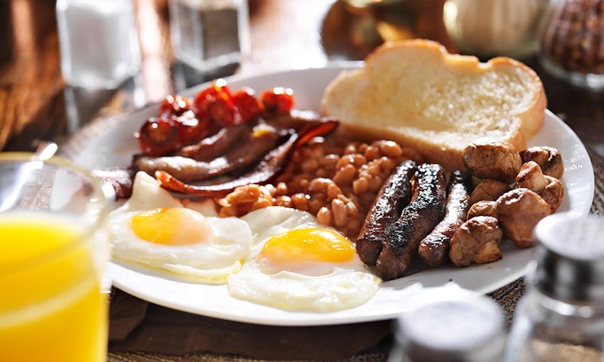 a full english breakfast consisting of a slice of white bread, fried mushrooms, three sausages, rashers of bacon, beans and two fried eggs with a glass of orange juice 