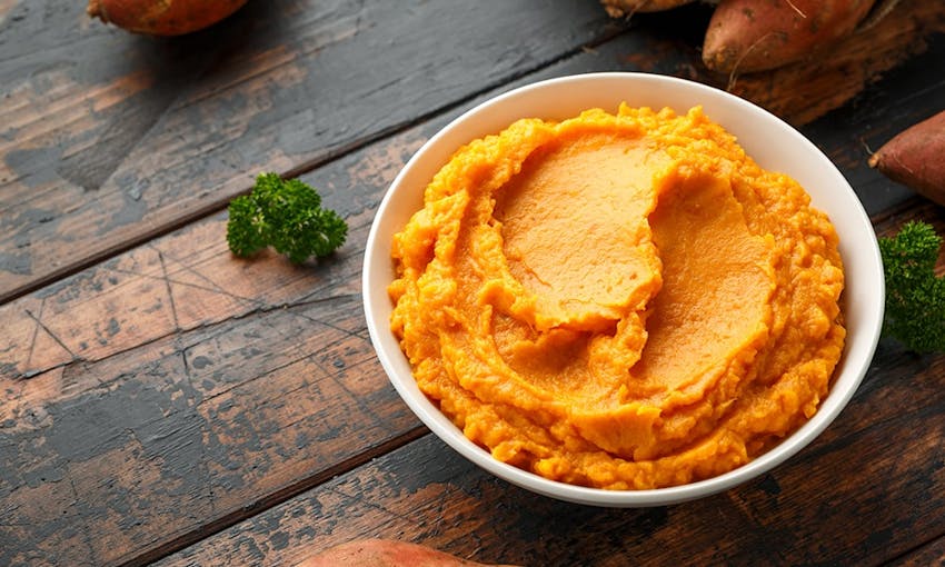 a bowl of sweet potato mash in a white bowl on a dark wooden table with sprigs of broccoli and next to whole sweet potatoes 