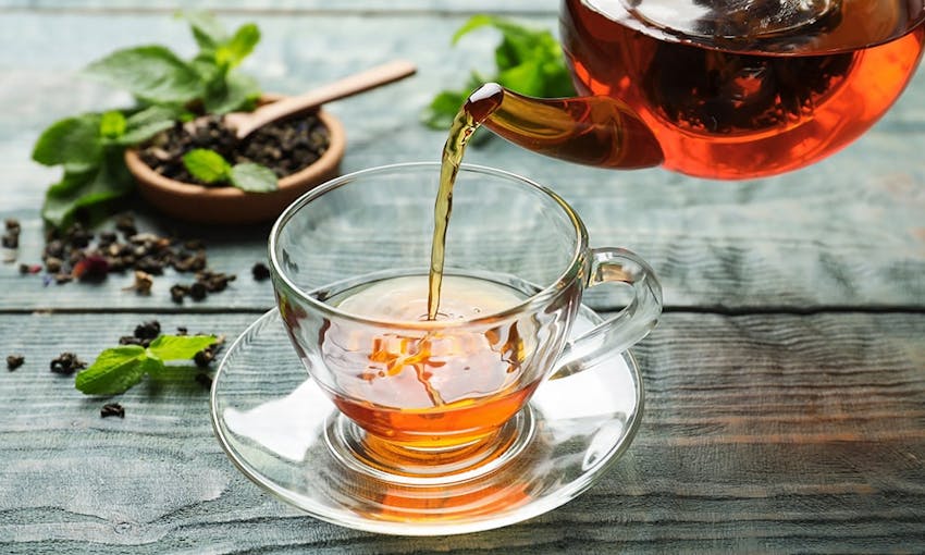 a clear glass teacup and saucer with herbal tea being poured into it on top of a wooden table with herbs and tea leaves scattered to the left 