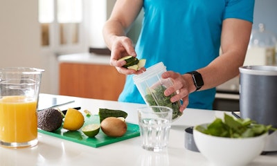 man in a blue tshirt adding healthy foods to a smoothie blender with some chopped fruits on chopping board and a glass of orange juice 