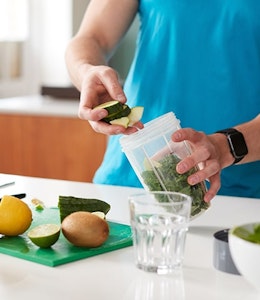 man in a blue tshirt adding healthy foods to a smoothie blender with some chopped fruits on chopping board and a glass of orange juice 