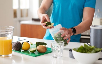 man in a blue tshirt adding healthy foods to a smoothie blender with some chopped fruits on chopping board and a glass of orange juice 