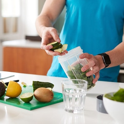 man in a blue tshirt adding healthy foods to a smoothie blender with some chopped fruits on chopping board and a glass of orange juice 