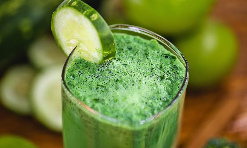 a forest green smoothie in a clear glass with a sliced garnish of cucumber 