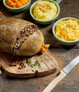 Haggis slightly chopped to reveal inside on a wooden chopping board surrounded by mini serving dishes of crispy onions, neeps and tatties  