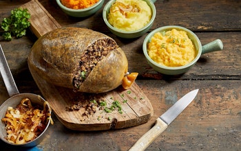 Haggis slightly chopped to reveal inside on a wooden chopping board surrounded by mini serving dishes of crispy onions, neeps and tatties  