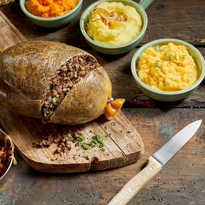 Burns Night Fun Facts Haggis slightly chopped to reveal inside on a wooden chopping board surrounded by mini serving dishes of crispy onions, neeps and tatties