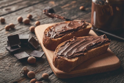 two thick slices of brown bread with a good helping of nutella on a wooden chopping board with dark chocolate pieces and hazelnuts to decorate the table 