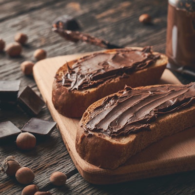 two thick slices of brown bread with a good helping of nutella on a wooden chopping board with dark chocolate pieces and hazelnuts to decorate the table 