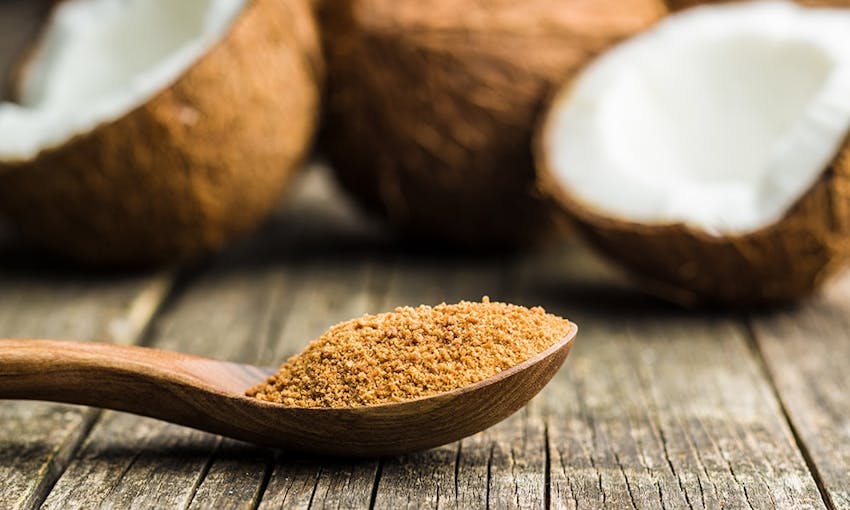 A teaspoon with coconut sugar on a wooden table in front of halved coconuts 