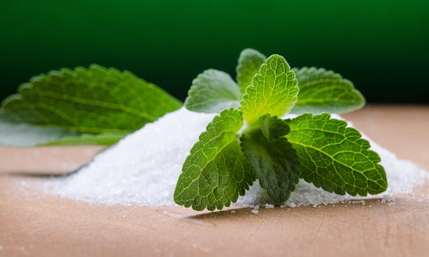 A small mound of stevia on wooden table with stevia leaf placed over the top 
