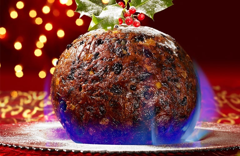 a round flaming christmas pudding on a silver platter on a decorated christmas christmas table with fairy lights in the background 