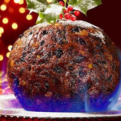 a round flaming christmas pudding on a silver platter on a decorated christmas christmas table with fairy lights in the background 
