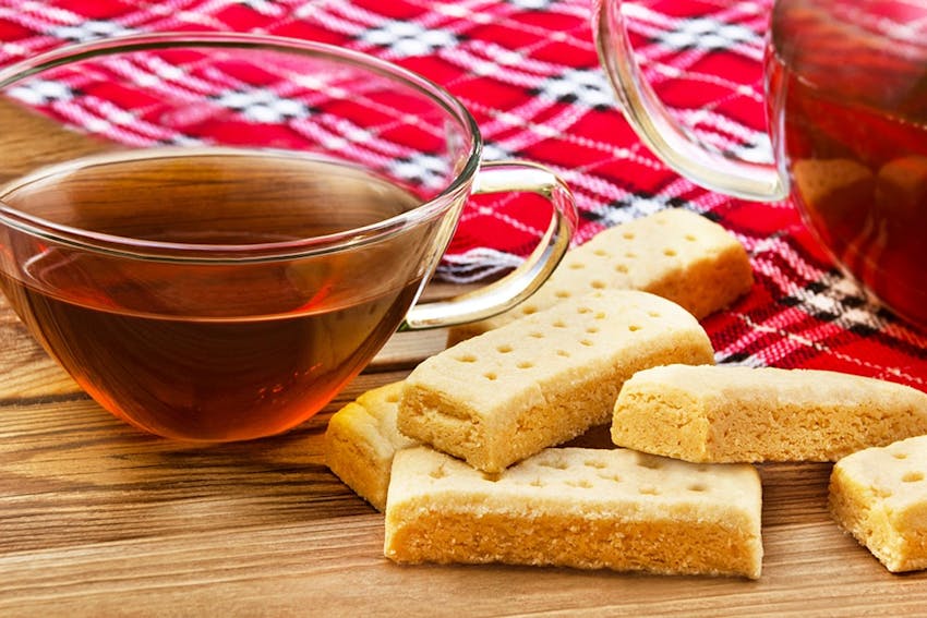 a cup of tea in a clear glass cup next to 6 shortbread fingers laid over a red tartan blanket 