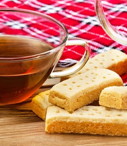 a cup of tea in a clear glass cup next to 6 shortbread fingers laid over a red tartan blanket 