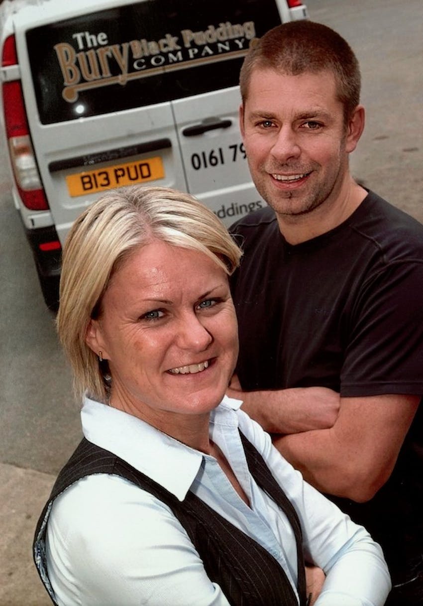 a man and woman posing arms crossed in front of a bury black pudding company van 