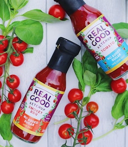 Real Good Ketchup's USP is their No Added Sugar  Two bottles of real good ketchup on a white wooden table with tomato plant and vine tomatoes for decoration