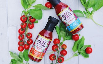 Two bottles of real good ketchup on a white wooden table with tomato plant and vine tomatoes for decoration 