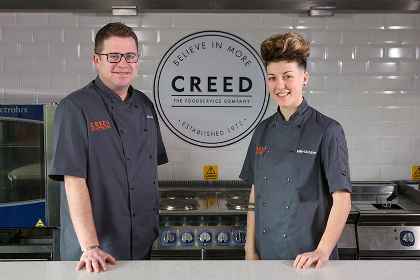 Male and female stood in creed  innovation centre in front of creed 'believe in more' logo 