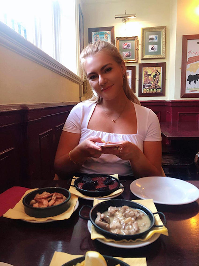 a young woman wearing a white tshirt and holding a mobile phone sits at a dinner table with three plates of tapas in front of her 