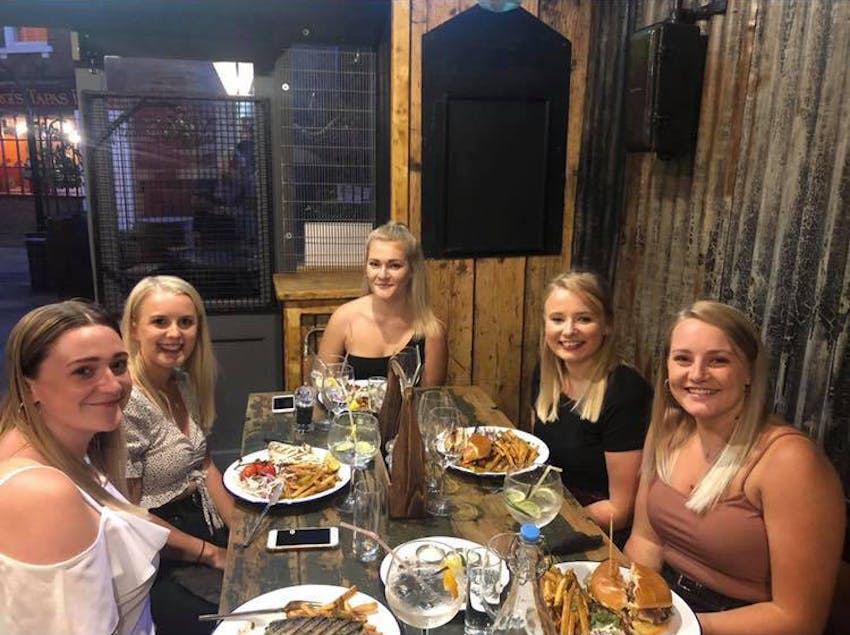 five girls sit around a wooden table in a restaurant smiling for the camera with plates full of food 