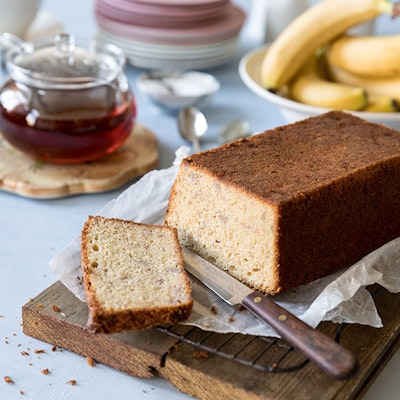baked banana bread cake with one slice cut off and a silver knife on top of a wooden chopping board 